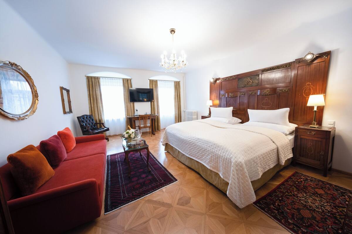 A hotel room with a large bed, a red sofa, a wooden headboard, a chandelier, and a television.