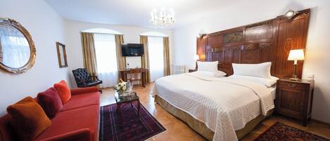 Habitación de hotel con una cama grande, un sofá rojo, un cabecero de madera, una araña y un televisor.