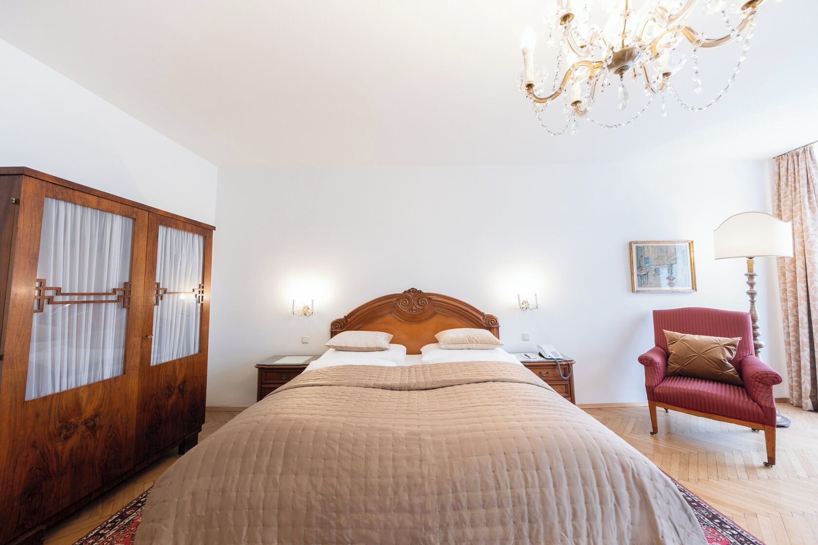 A bedroom with a large bed, wooden wardrobe, a red armchair, a chandelier, and a nightstand with a lamp.