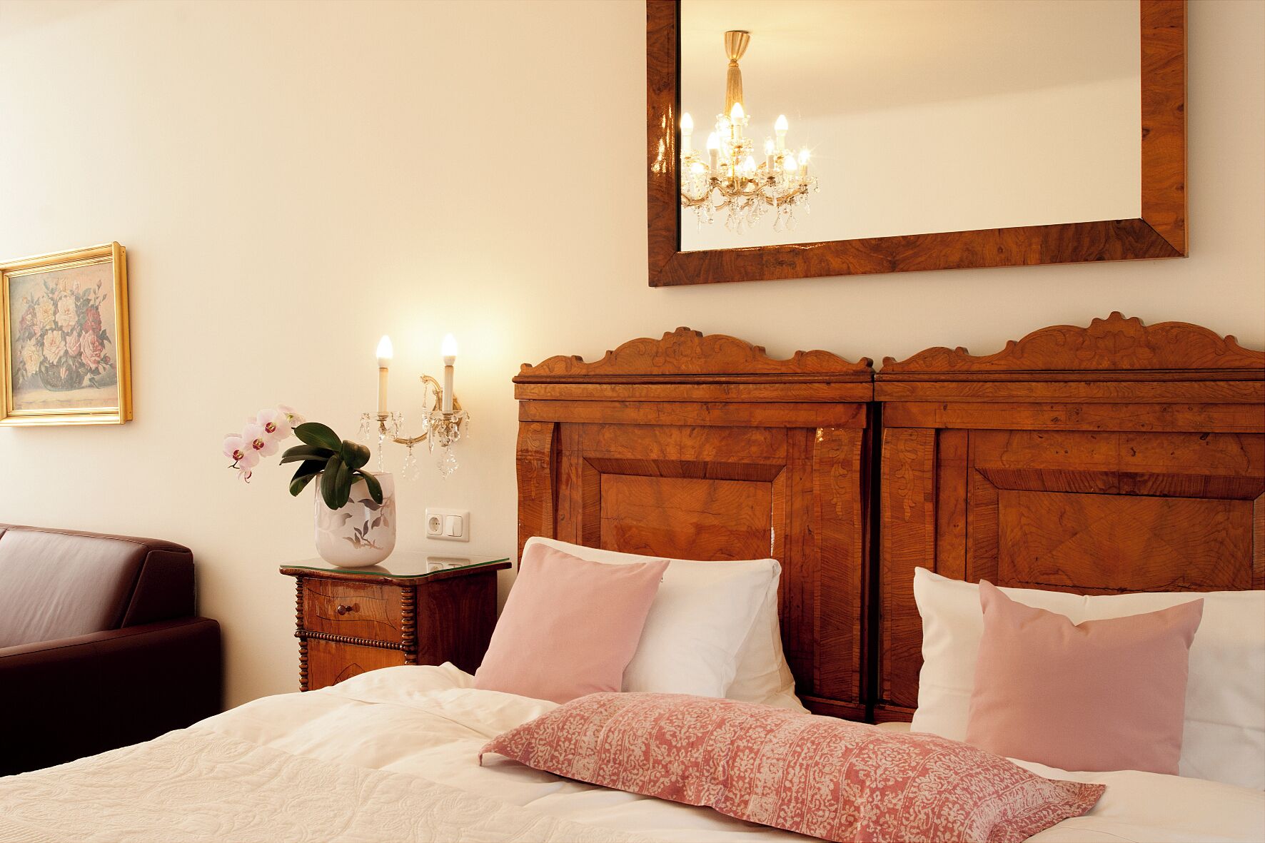 A bedroom with a wooden headboard, a bed with white and pink bedding, a nightstand with a vase of flowers, and a chandelier.