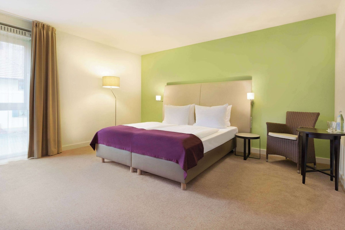 A hotel room with a bed, a chair, a small table, and a lamp.