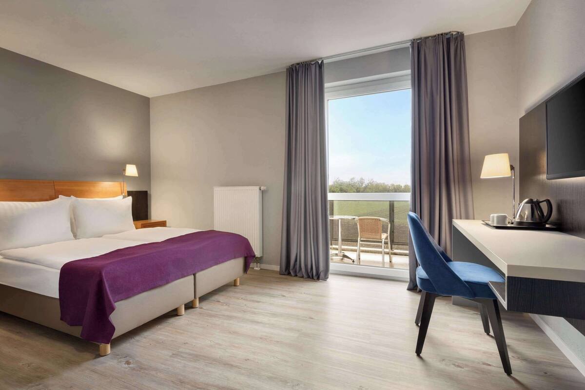A hotel room with a bed, a desk, a chair, a TV, and a balcony with curtains.