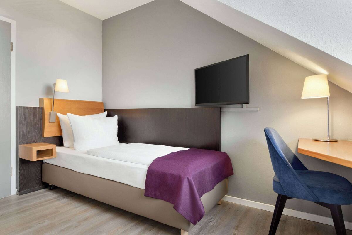 A modern hotel room with a bed, a desk, a chair, a TV, and a lamp.