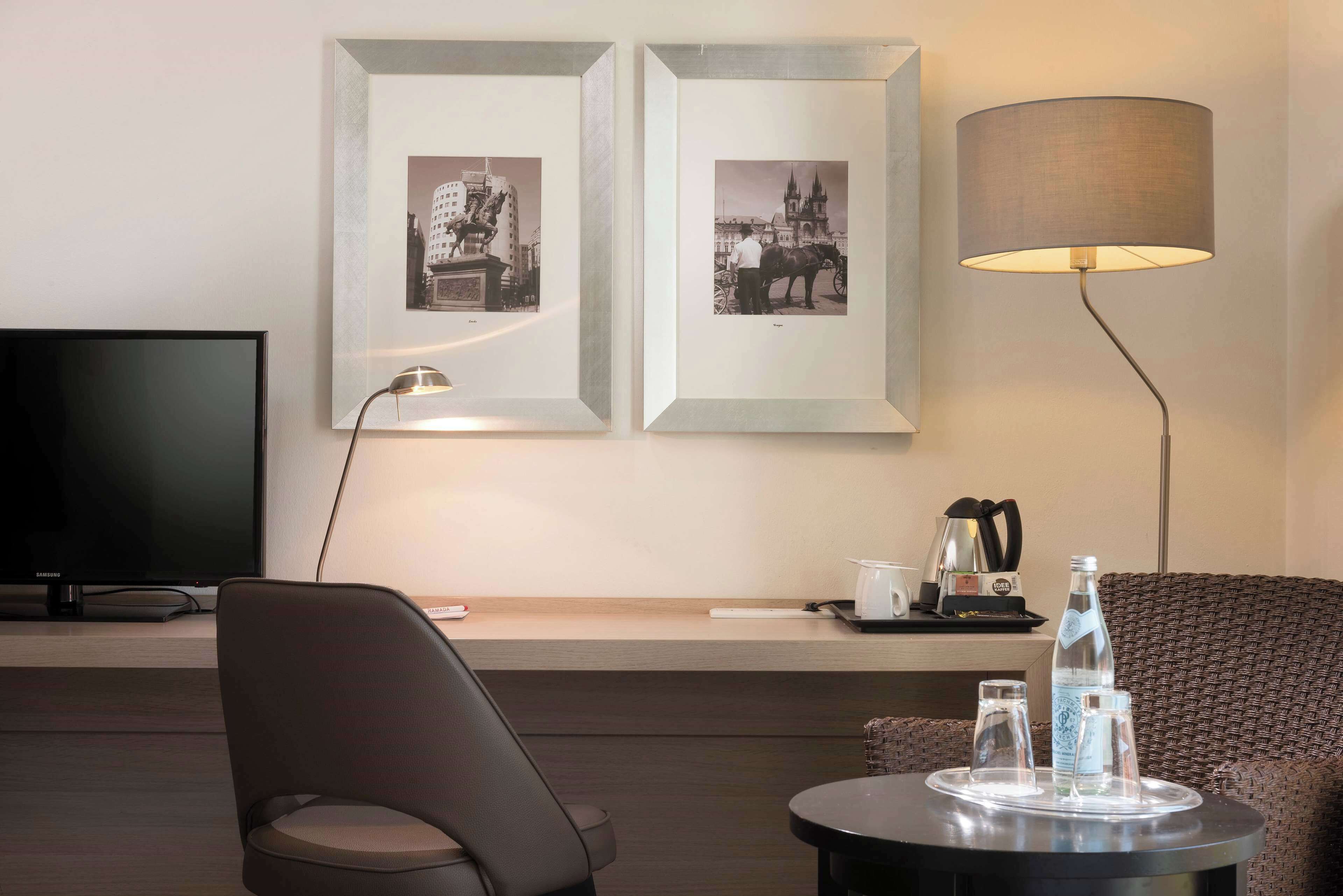 Een moderne hotelkamer met een flatscreen televisie, twee ingelijste zwart-wit foto’s aan de muur, een vloerlamp, een rieten bank, een klein rond tafeltje met glazen en een fles, en een dienblad met een koffiezetapparaat.