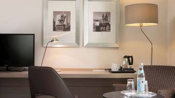 A modern hotel room with a flat-screen TV, two framed black and white photographs on the wall, a standing lamp, a wicker sofa, a small round table with glasses and a bottle, and a tray with a coffee maker.