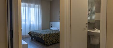 A hotel room with a bed, a bedside table, a window with curtains, and a bathroom with a sink and mirror.