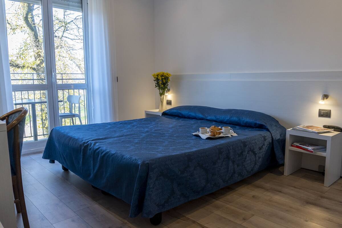 A bedroom with a large bed, a bedside table with a lamp, a chair, and a window with a view of trees.