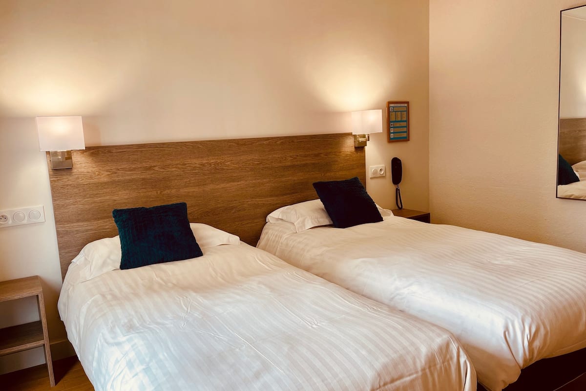 Habitación de hotel con dos camas, cada una con ropa de cama blanca y almohadas azul oscuro. Tiene un cabecero de madera, una mesita de noche con lámpara y un teléfono en la pared.