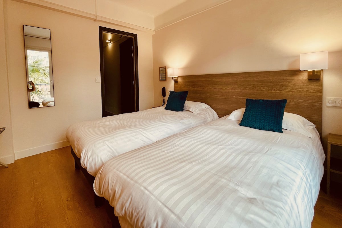 Habitación de hotel con dos camas, cabecero de madera, mesita de noche, espejo y lámpara fijada a la pared.