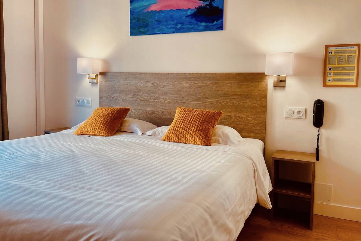 Una cama bien tendida con dos almohadas, un cabecero de madera, una mesita de noche con una lámpara y un cuadro en la pared.