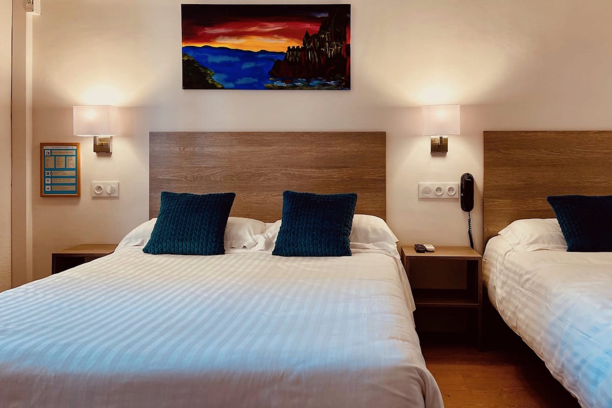 Habitación de hotel con dos camas, cada una con almohada azul oscuro y ropa de cama blanca. Hay lámparas fijadas a la pared, un cuadro de un paisaje y un cabecero de madera.