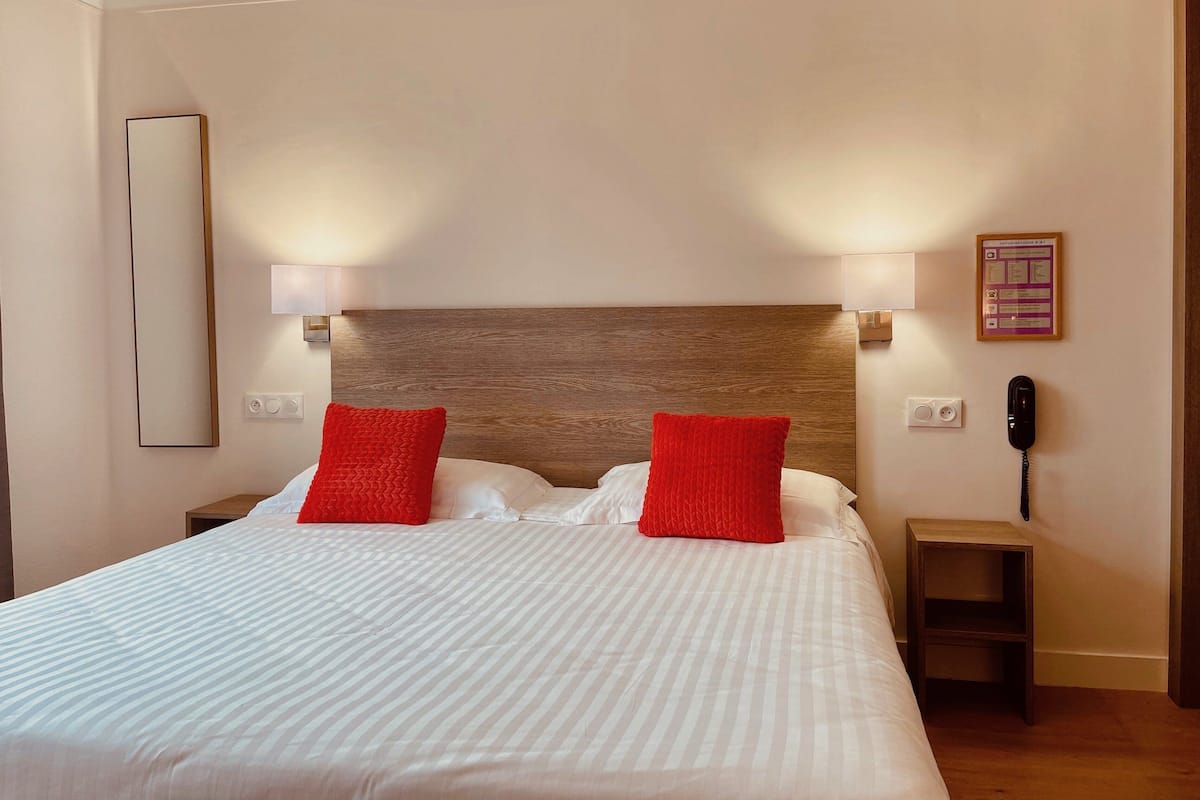 Una cama bien hecha con sábanas blancas y dos almohadas rojas, un cabecero de madera, lámparas de noche, una mesita de noche de madera y un teléfono instalado en la pared.