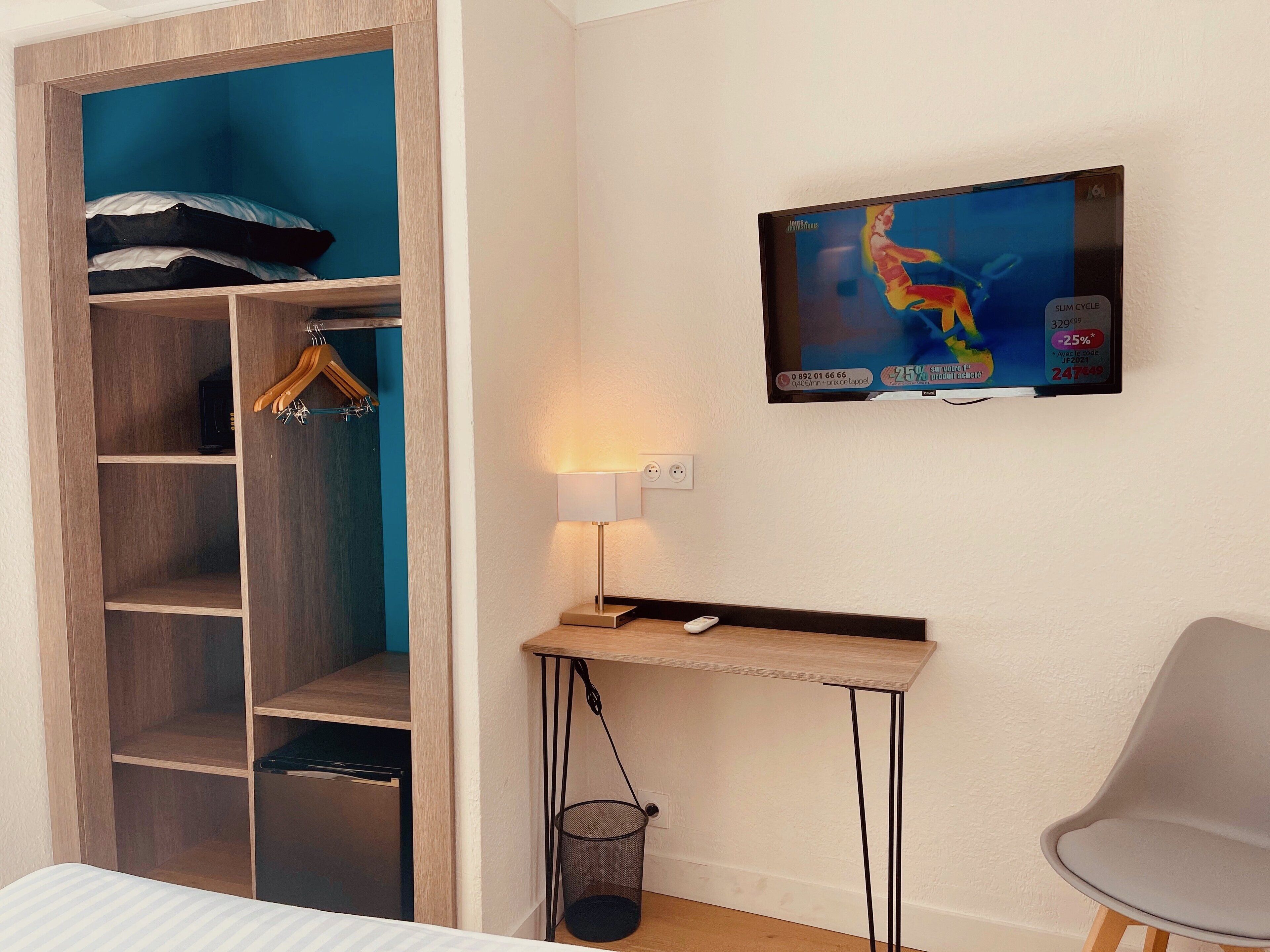 A hotel room with a wardrobe, a desk with a lamp, a TV on the wall, and a chair.