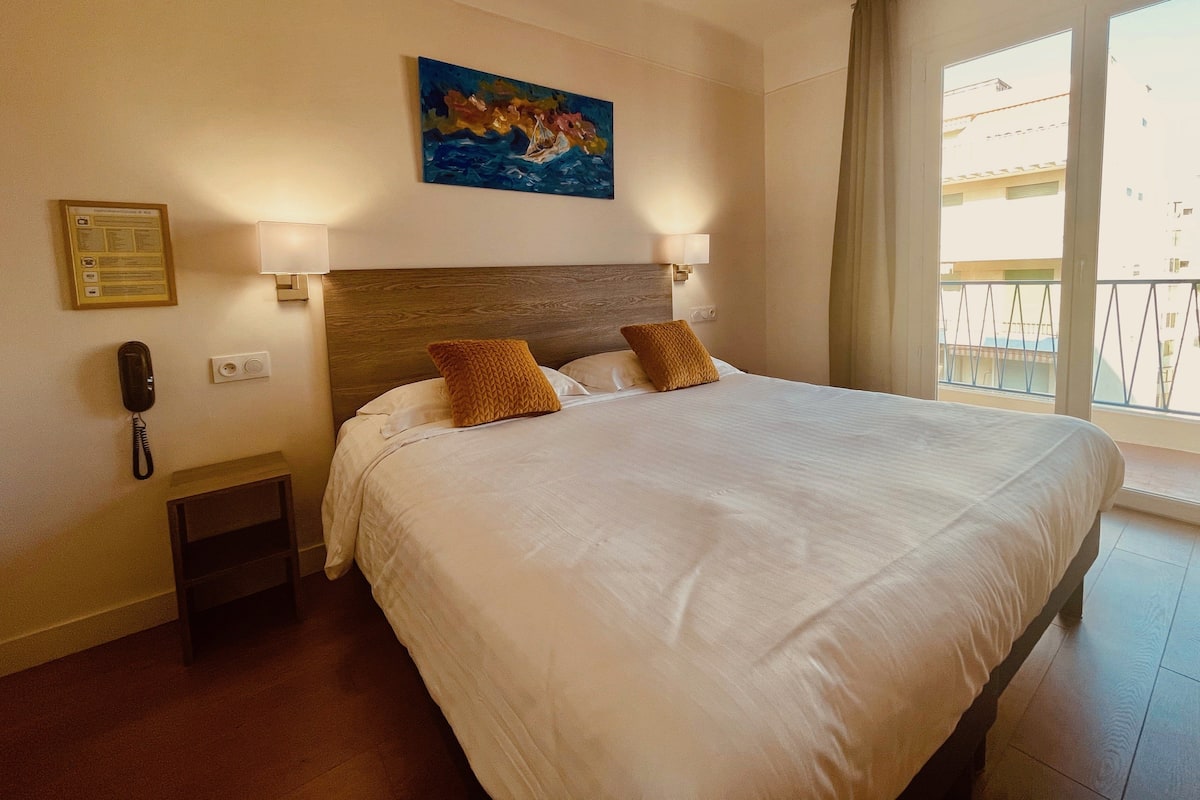 Habitación de hotel con una cama grande, dos almohadas, una mesita de noche con un teléfono, un cuadro en la pared y una ventana con vistas a edificios.