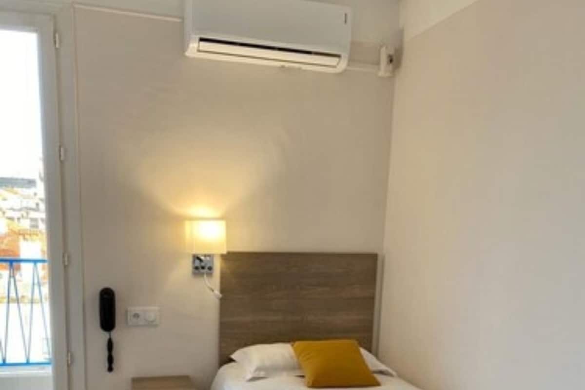 Un dormitorio con cama, mesita de noche, lámpara, aire acondicionado y puerta a un balcón.