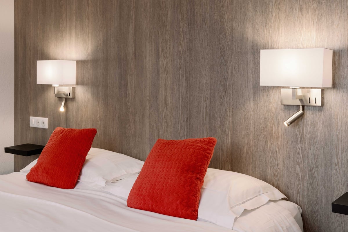 Habitación de hotel con cabecera de madera, dos mesitas de noche, ropa de cama blanca y almohadas rojas.