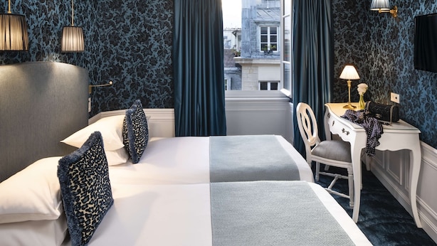 A hotel room with two beds, a desk, a chair, a lamp, and a view of the city through the window. - Odéon Hôtel  (Paris)