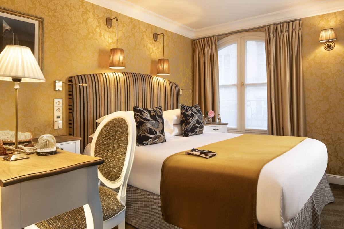 A hotel room with a large bed, a desk with a lamp, a chair, and a window with curtains.