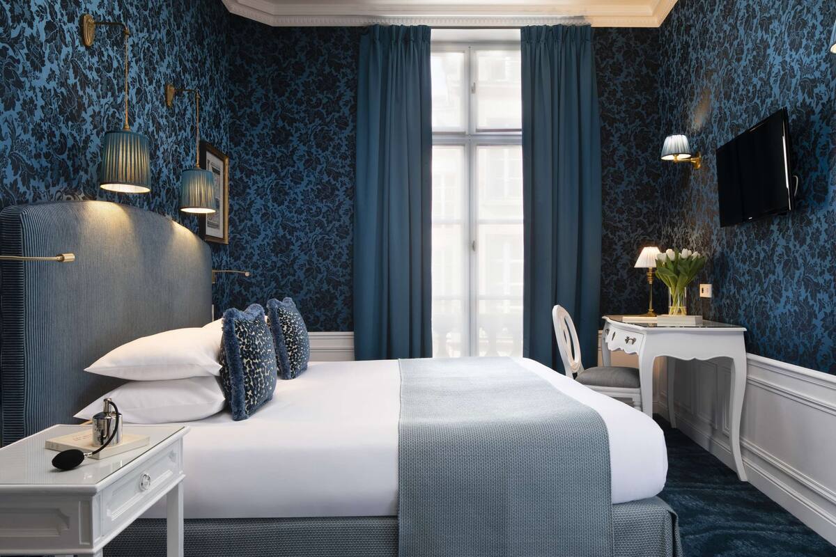 A hotel room with a large bed, a desk with a lamp, a TV, and a window with curtains.