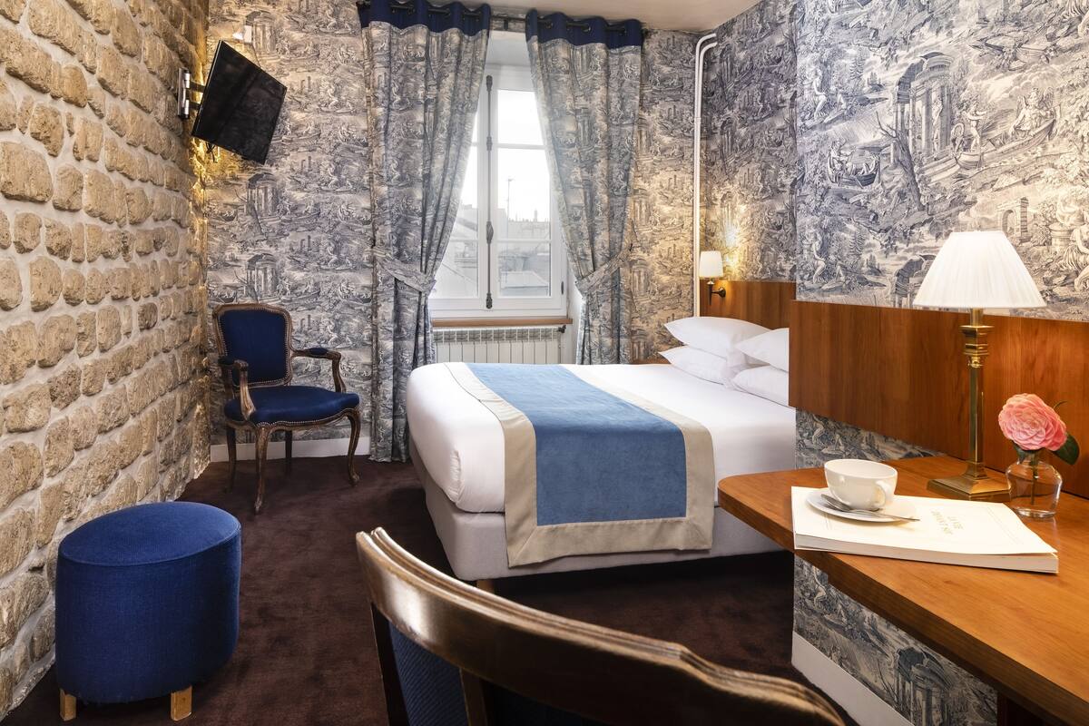 A hotel room with a stone wall, a bed with a blue blanket, a wooden desk with a lamp, a chair, and a window with curtains.