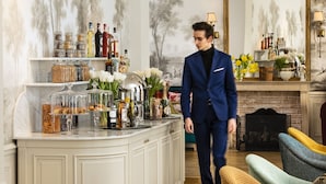 A man in a blue suit in a room with a white cabinet filled with bottles and jars, a fireplace, and a mirror. - Odéon Hôtel  (Paris)