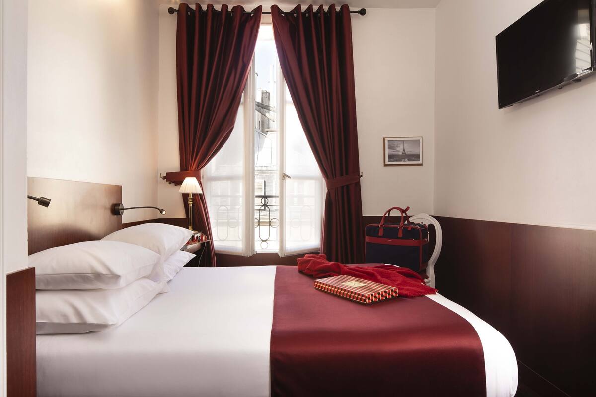 A hotel room with a bed, a window with curtains, a television on the wall, and a suitcase on a chair.