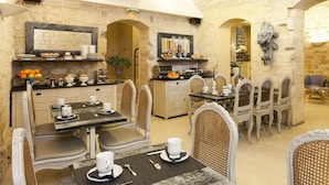 A dining area with a stone wall, a buffet with fruits and pastries, and a seating arrangement with white upholstered chairs. - Hôtel Ducs D'Anjou (Paris)