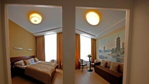 Hotellihuone, jossa on sänky, oleskelualue pöydällä ja tuoleilla sekä suuri ikkuna, jossa on verhot.