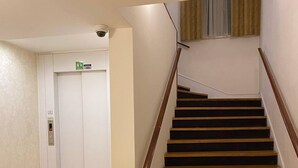 Portaat, joissa on puiset askelmat ja kaide, johtavat hissiin, jonka yläpuolella on vihreä hätäuloskäyntiä osoittava kyltti.