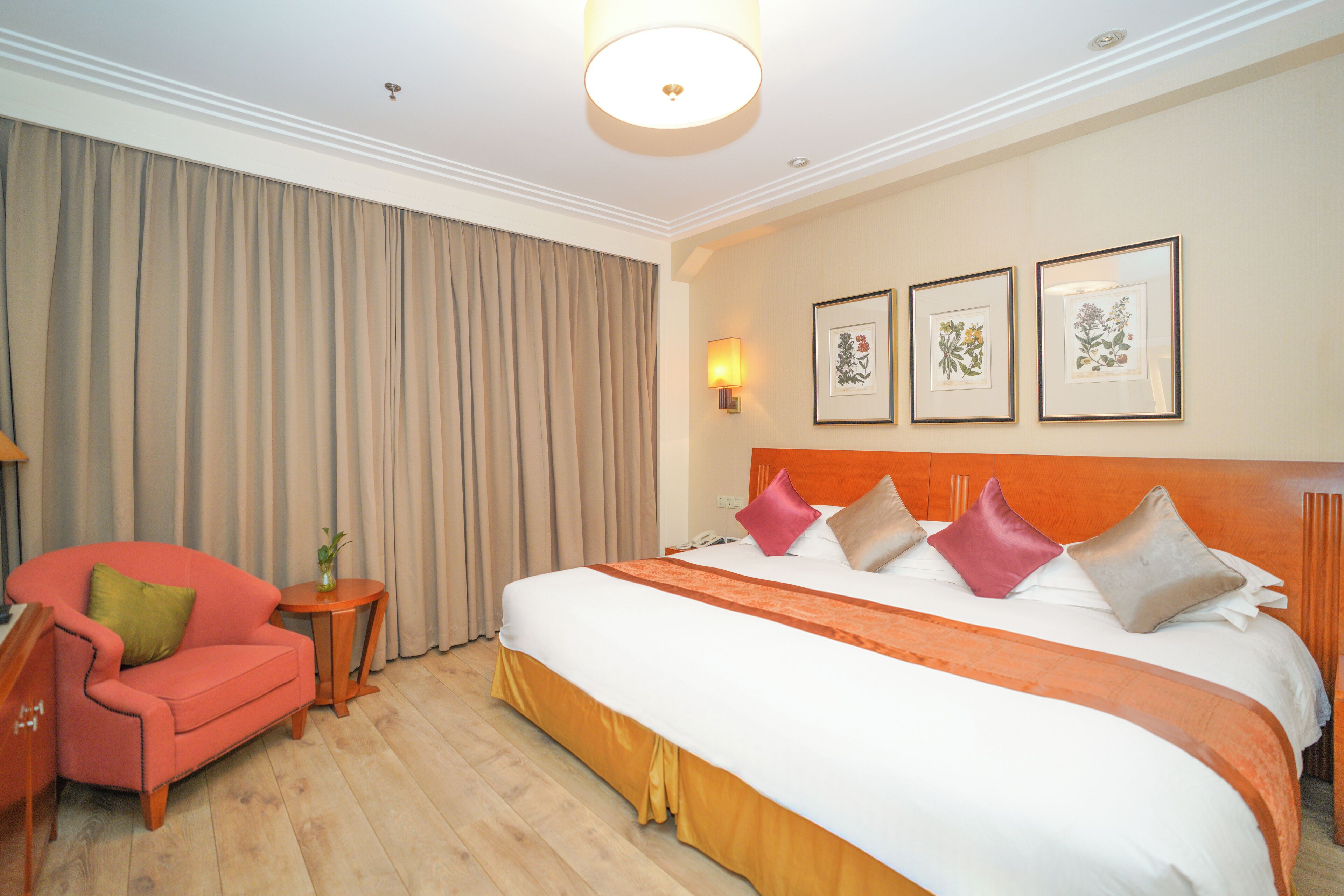 A hotel room with a large bed, a red armchair, a small table with a vase, and three framed botanical prints on the wall.