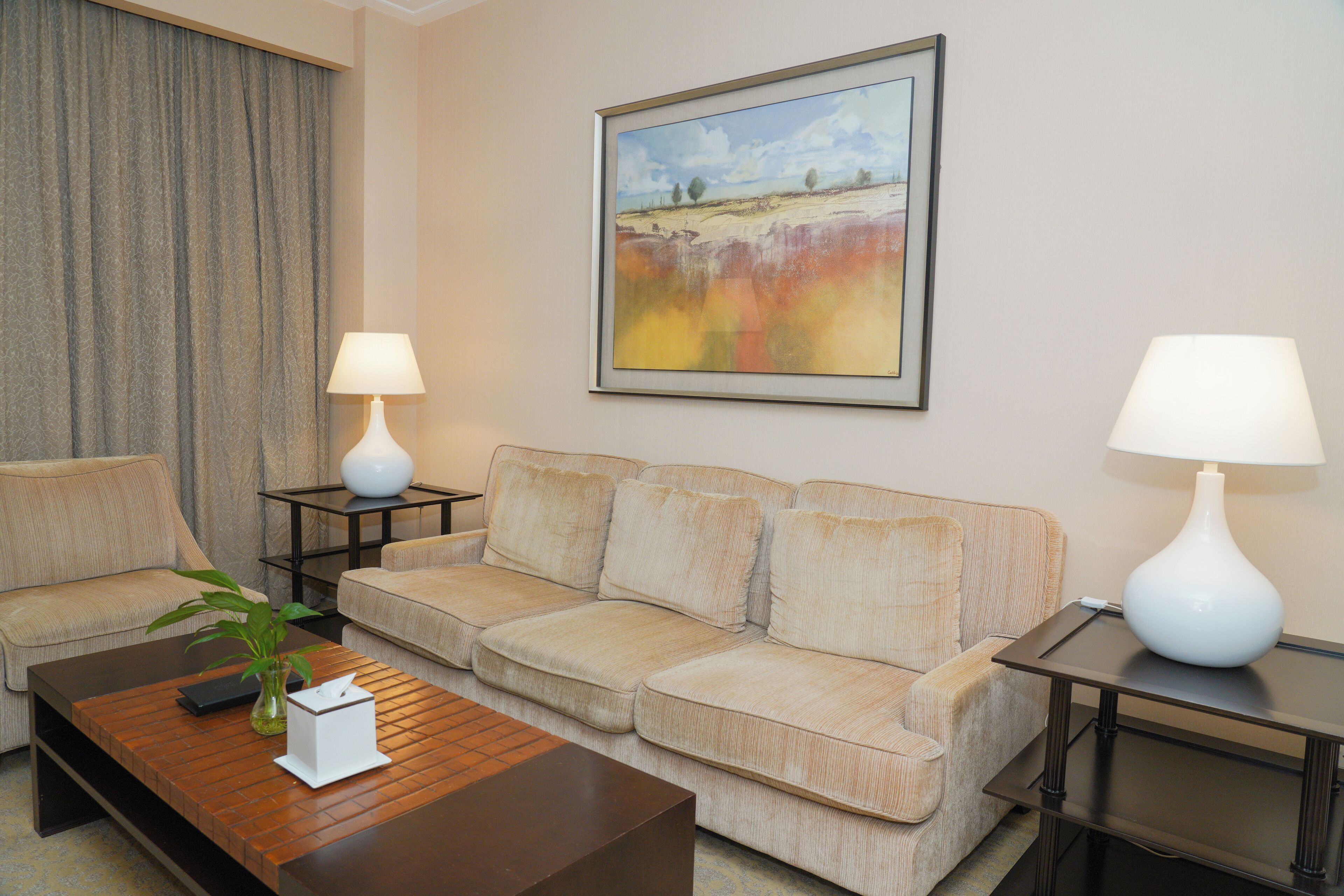 Una sala de estar con un sofá color beige, dos lámparas blancas, un cuadro enmarcado y una mesa de centro con una planta.