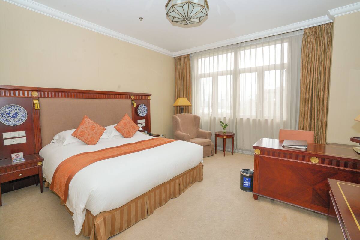 Quarto de hotel com uma cama grande, uma escrivaninha, uma cadeira e uma mesinha de cabeceira com abajur.