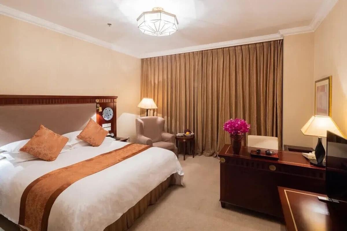 Quarto de hotel com uma cama grande, uma escrivaninha, uma cadeira, um abajur, um relógio e uma televisão.