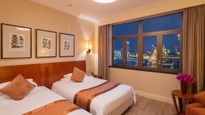 Habitación de hotel con dos camas, una mesita de noche, una lámpara, una ventana con vistas a la ciudad e impresiones botánicas enmarcadas en la pared.