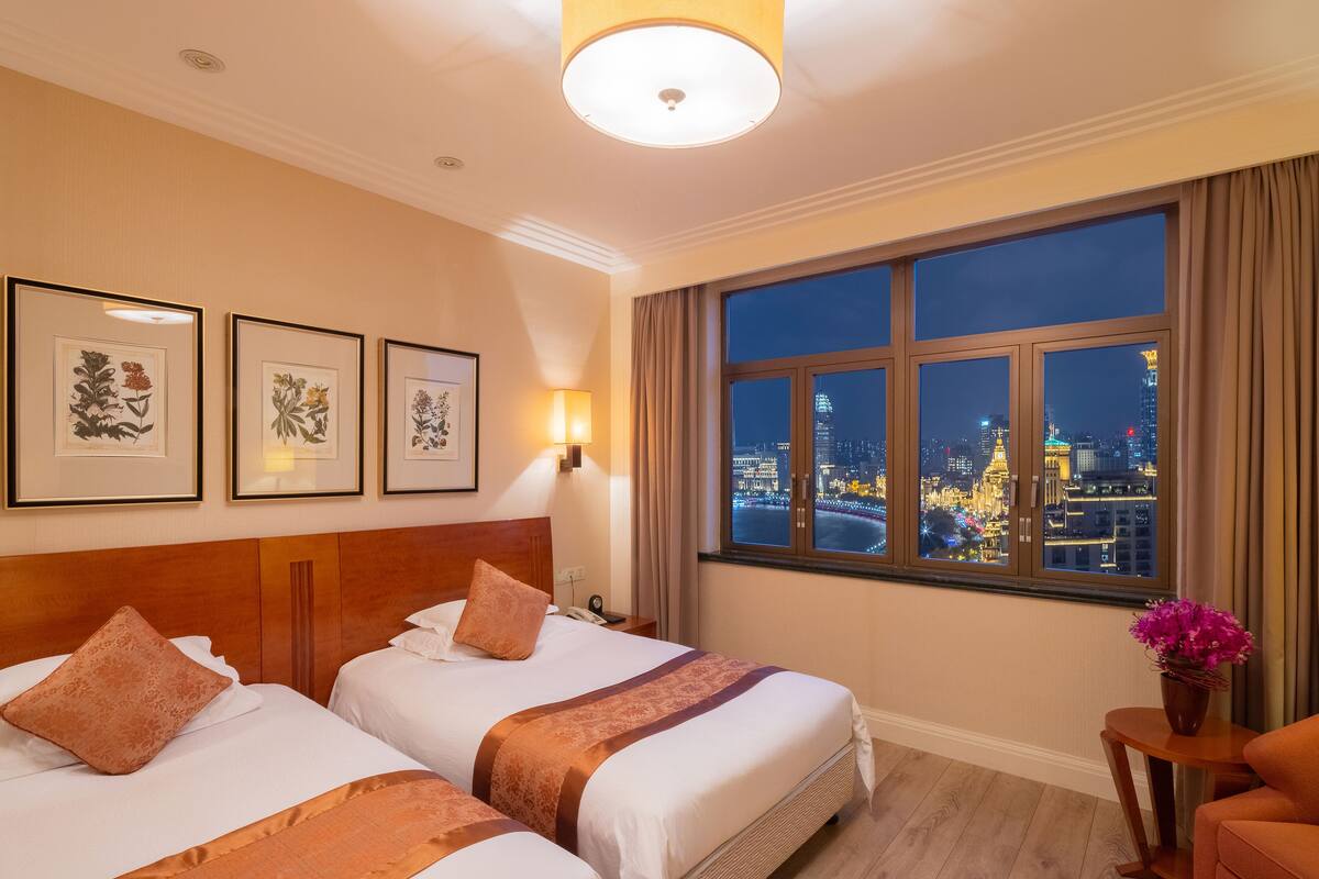 Quarto de hotel com duas camas, uma mesa de cabeceira, uma luminária, uma janela com vista para a cidade e impressões botânicas emolduradas na parede.