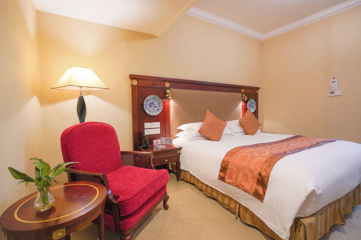 Quarto de hotel com cama, poltrona vermelha, mesa de cabeceira de madeira, abajur e vaso com planta.