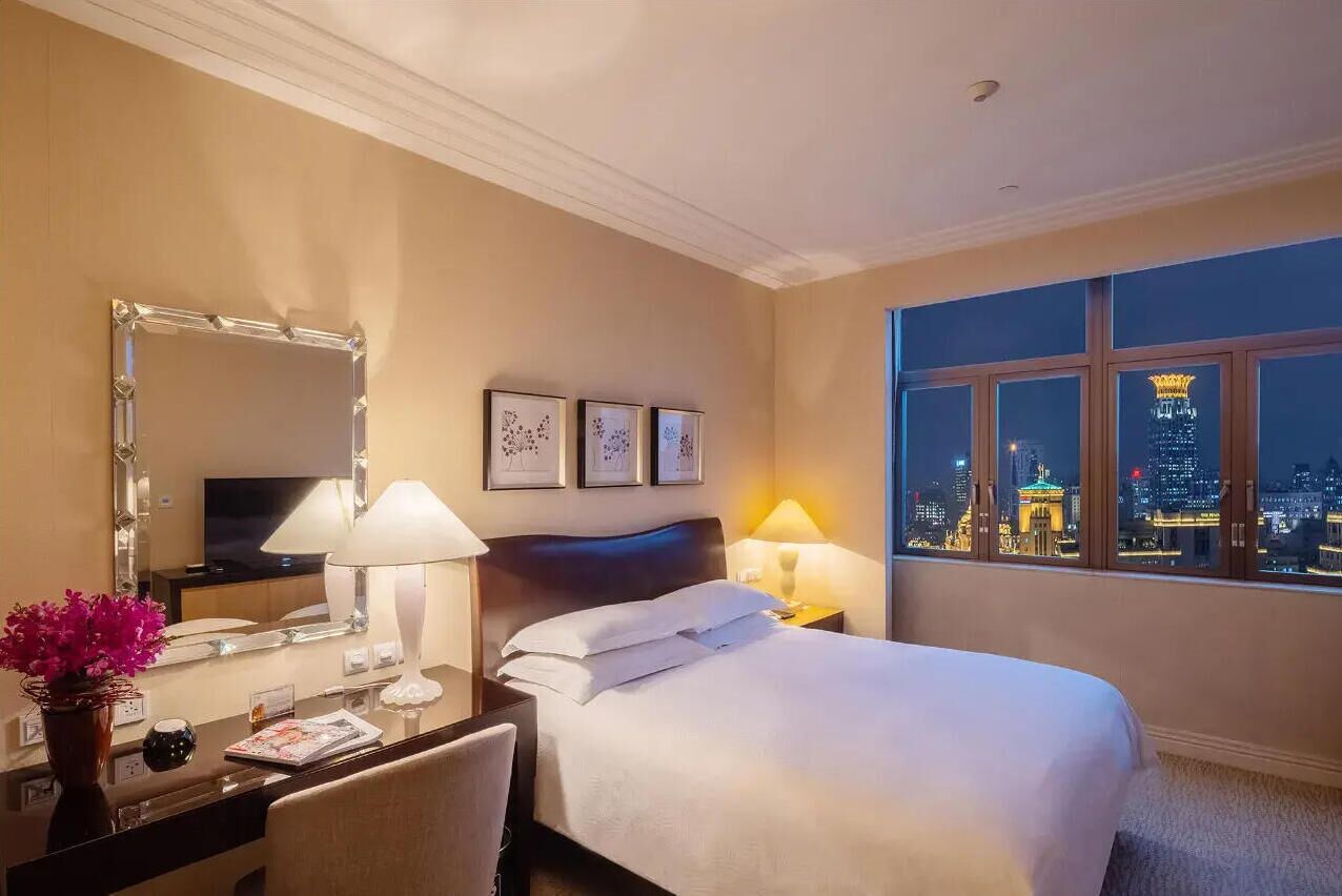 Ein Hotelzimmer mit einem Bett, Nachttischlampen, einem Schreibtisch mit Lampe, einem Spiegel und gerahmten Bildern an der Wand.
