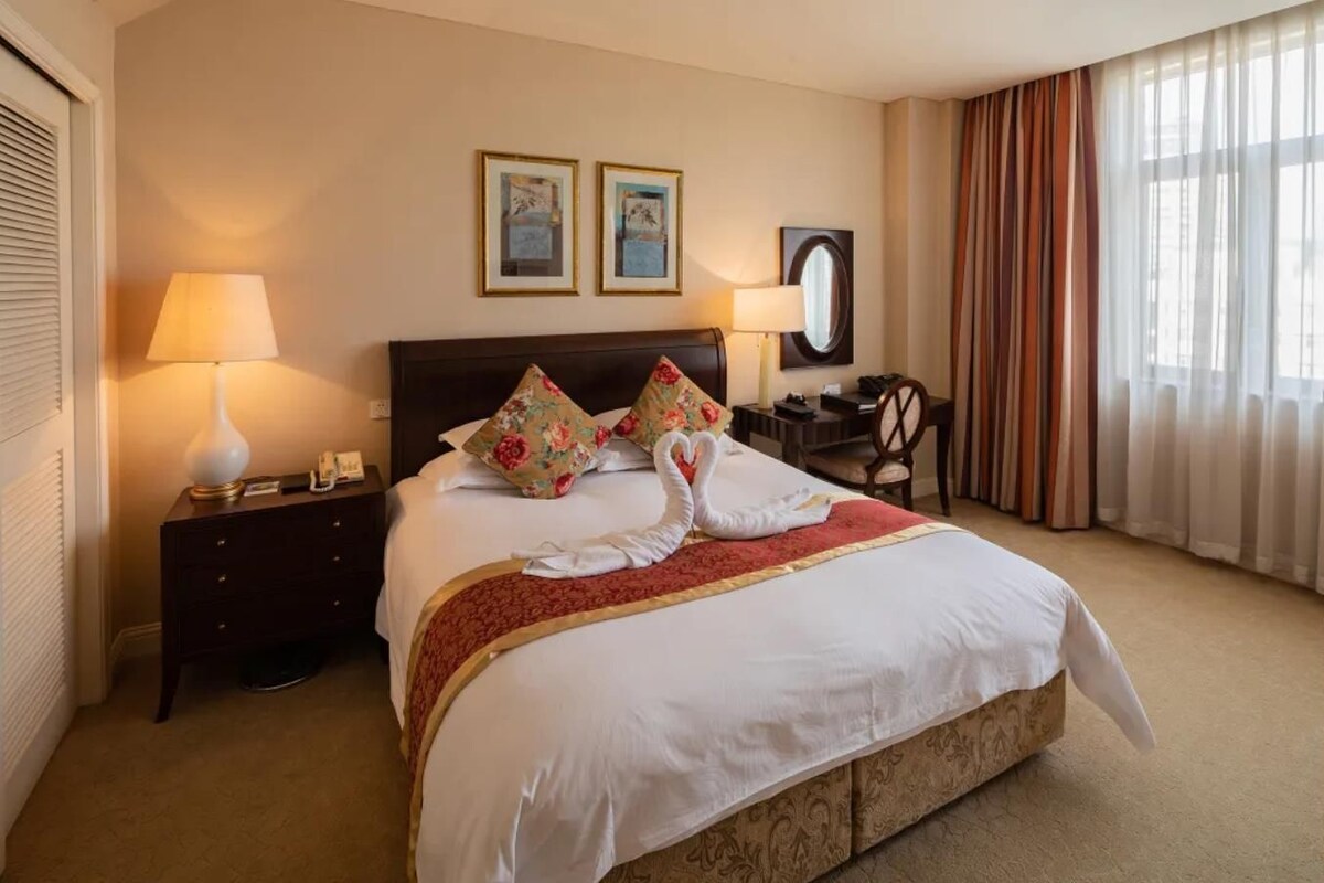 A hotel room with a large bed, two bedside lamps, a desk with a chair, a mirror, and two framed pictures on the wall.