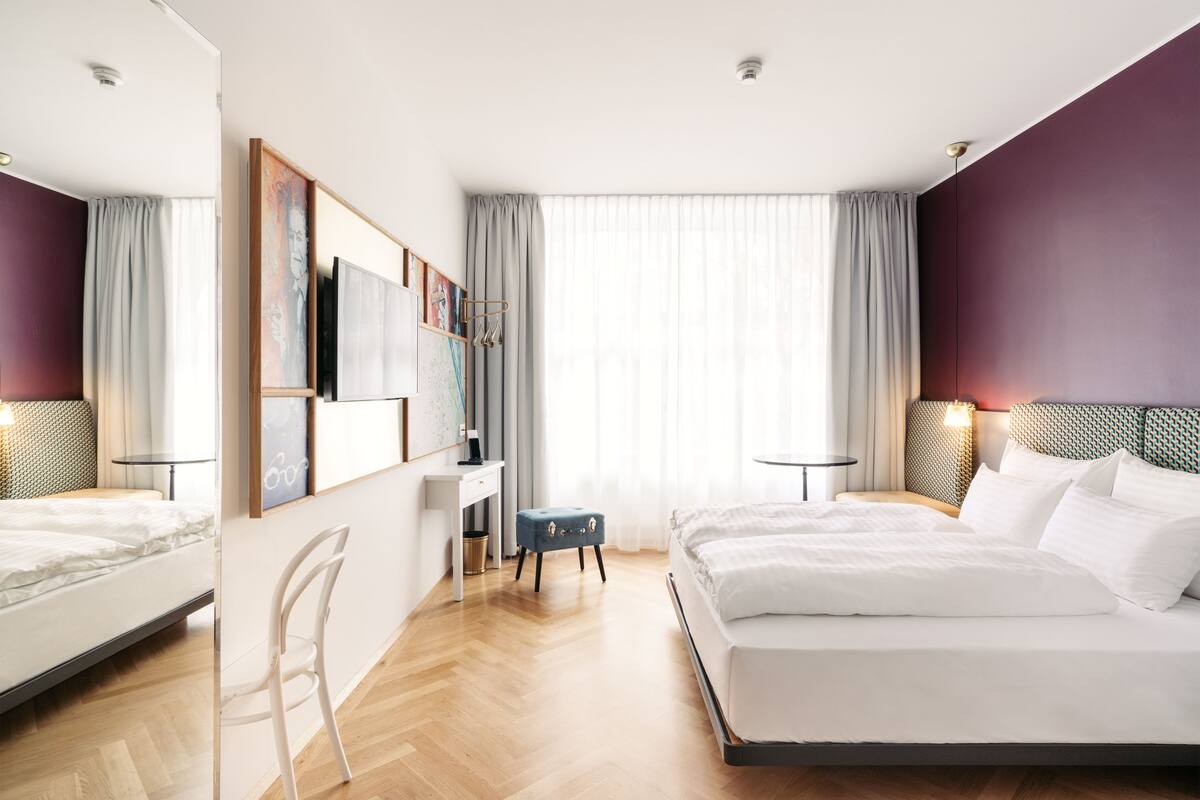 A modern hotel room with a large bed, a desk, and a chair. There is a window with curtains, a painting on the wall, and a lamp on the desk.