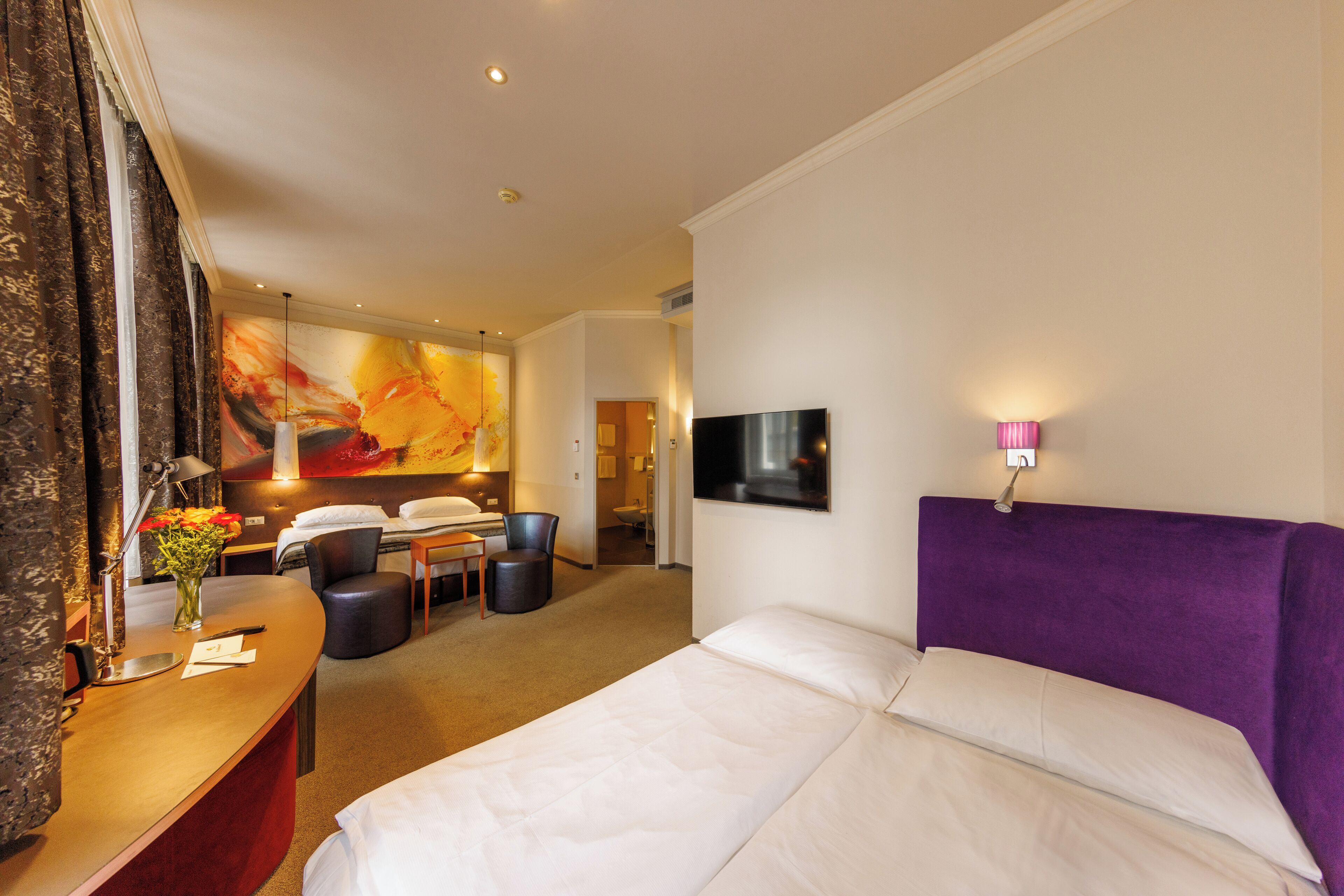 Een hotelkamer met een groot bed, een bureau met lamp, een stoel en een televisie aan de muur.