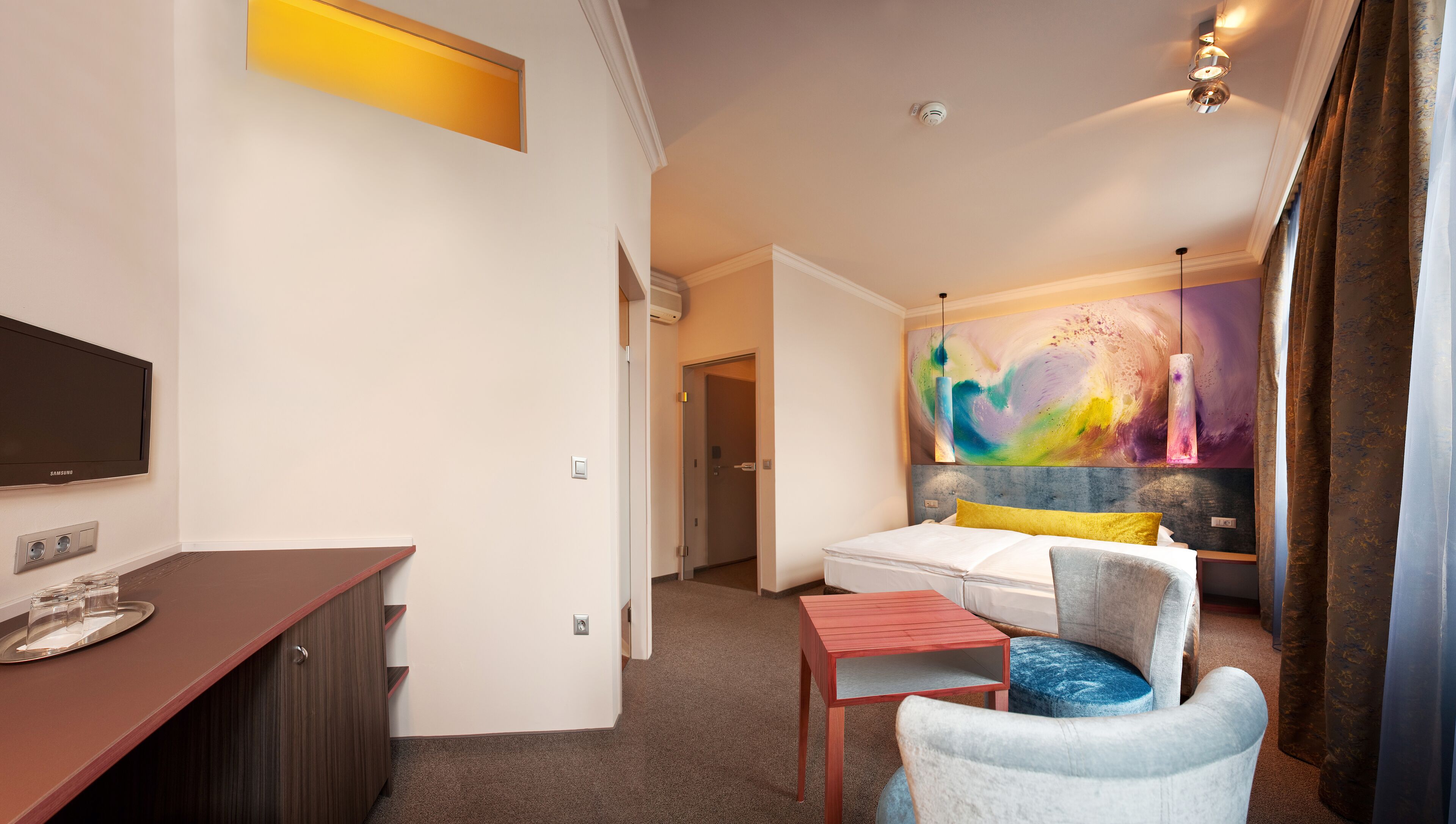 Een hotelkamer met een bed, een bureau met een televisie, een schilderij aan de muur en een raam met gordijnen.