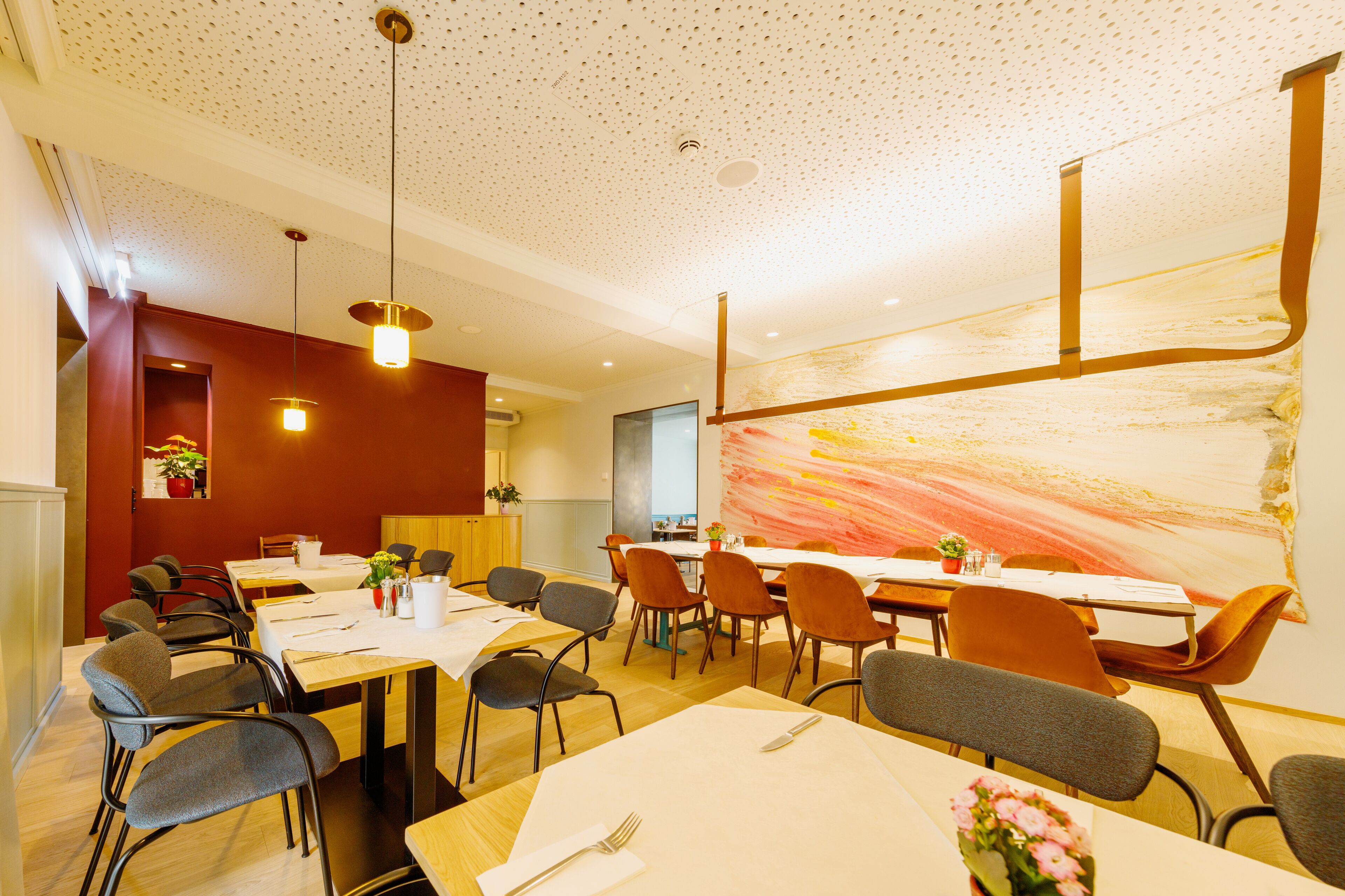 A dining area with multiple tables and chairs, a ceiling with a perforated pattern, and a large abstract painting on the wall.