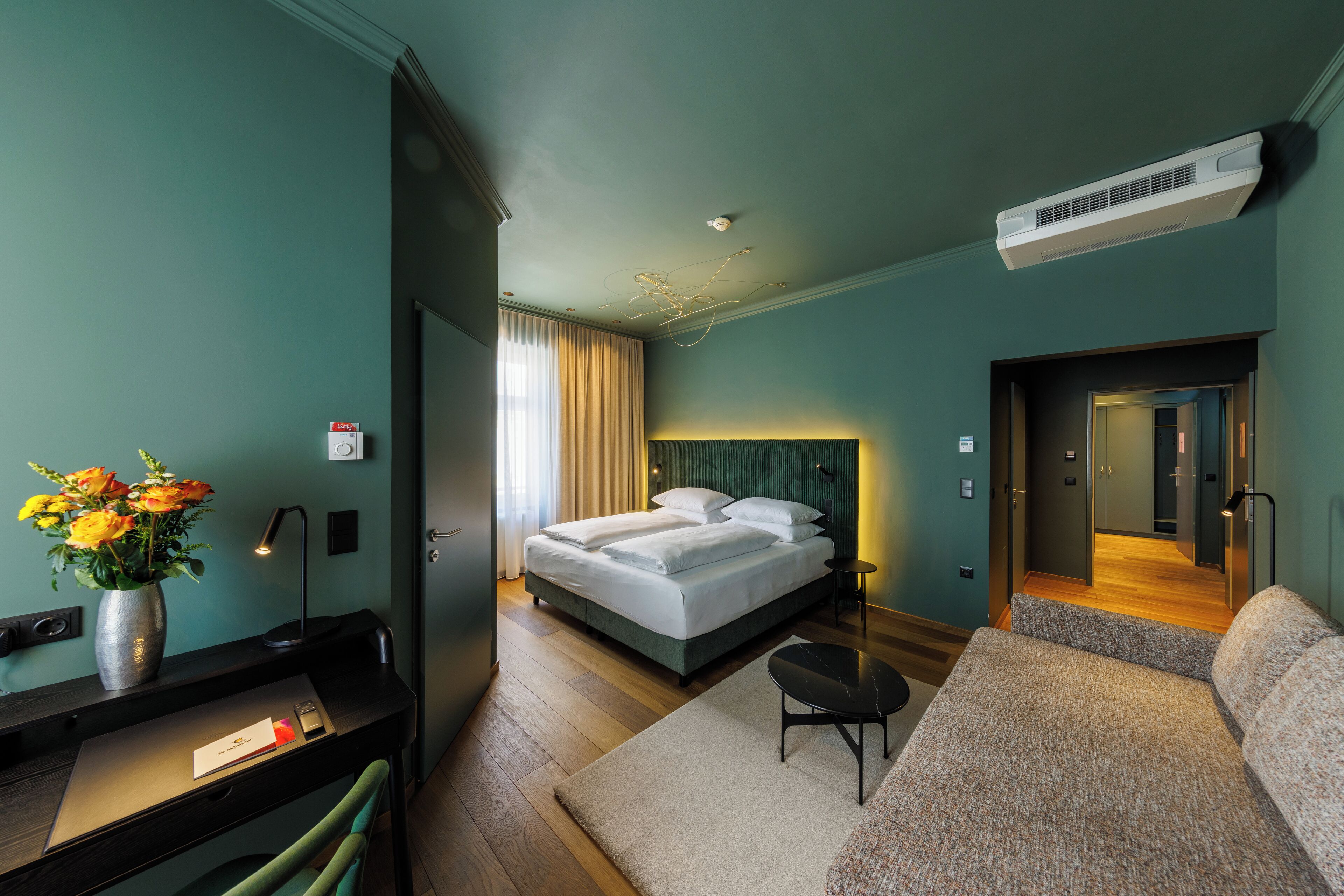 A modern hotel room with a bed, a desk, a chair, a sofa, and a vase of flowers.