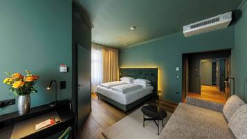 A modern hotel room with a bed, a desk, a chair, a sofa, and a vase of flowers.