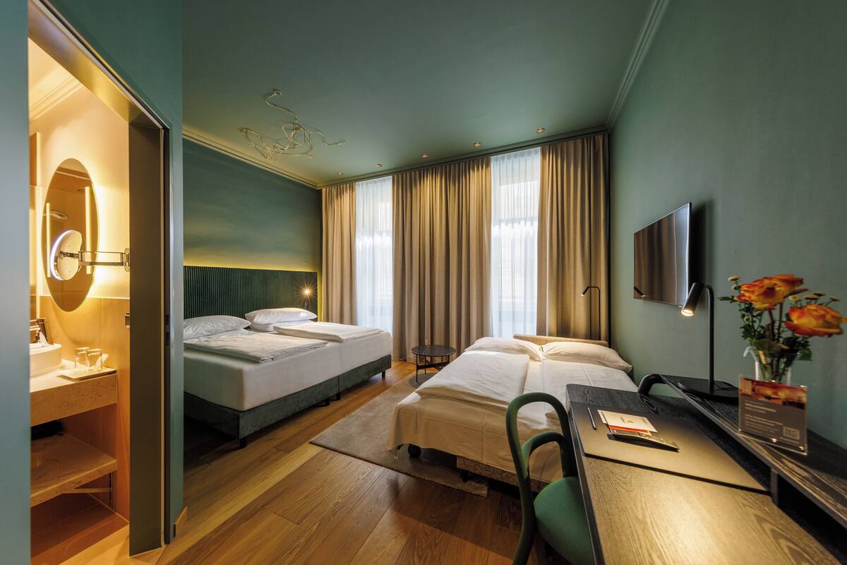 A hotel room with two beds, a desk, a chair, a TV, and a bathroom with a mirror and sink.