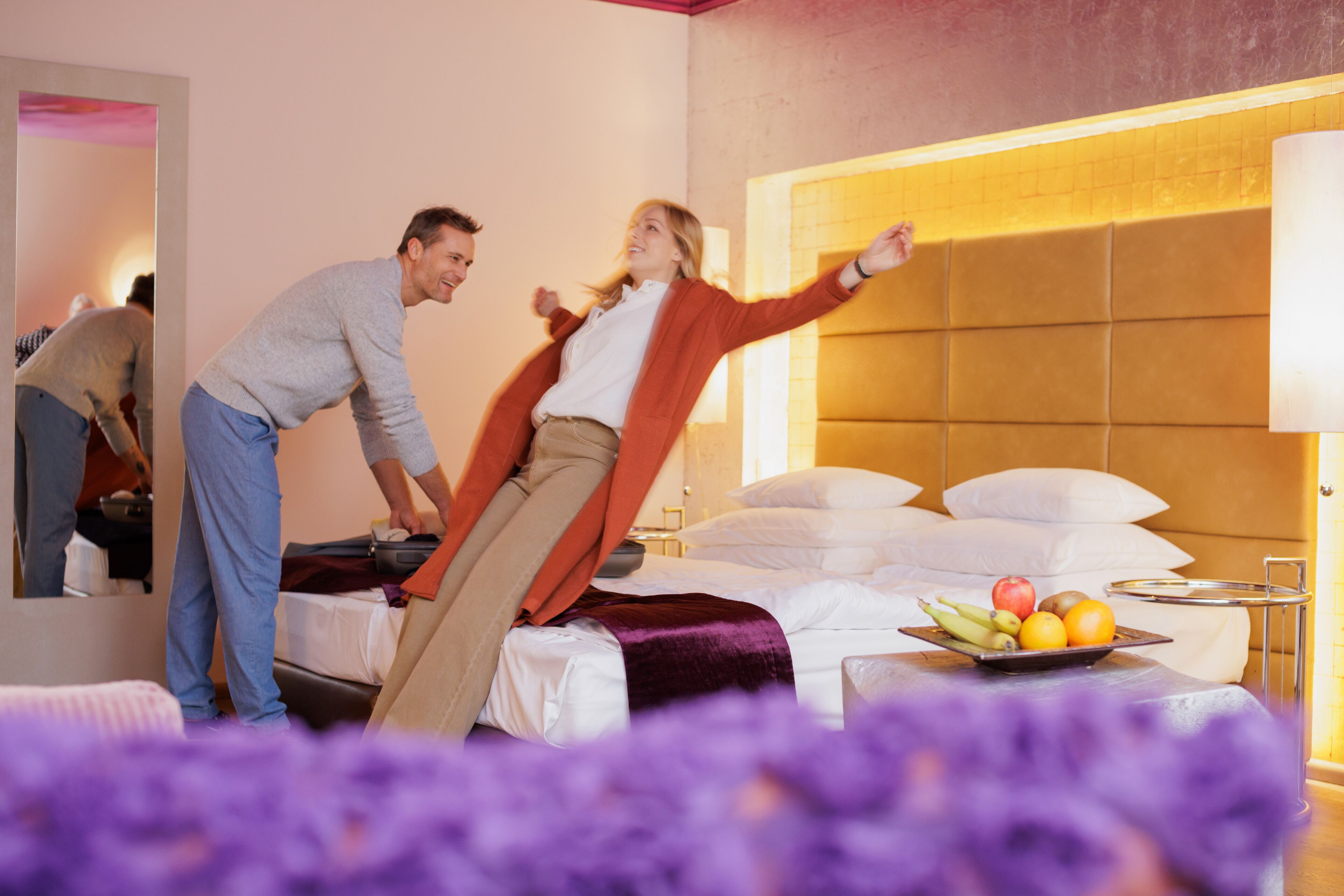 A hotel room with a bed, a bedside table with fruit, and a mirror reflecting part of the room.