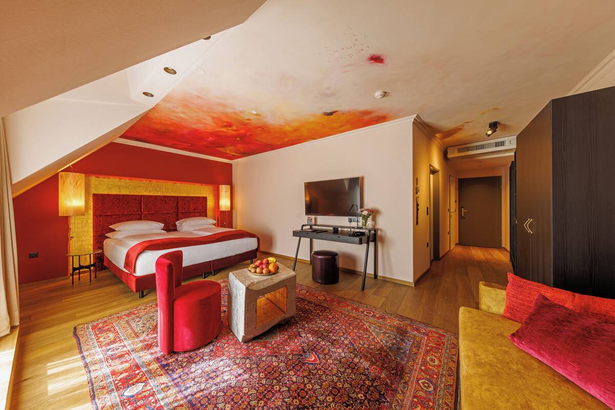 A modern hotel room with a large bed, a TV, a small table with fruit, a red chair, and a colorful rug.
