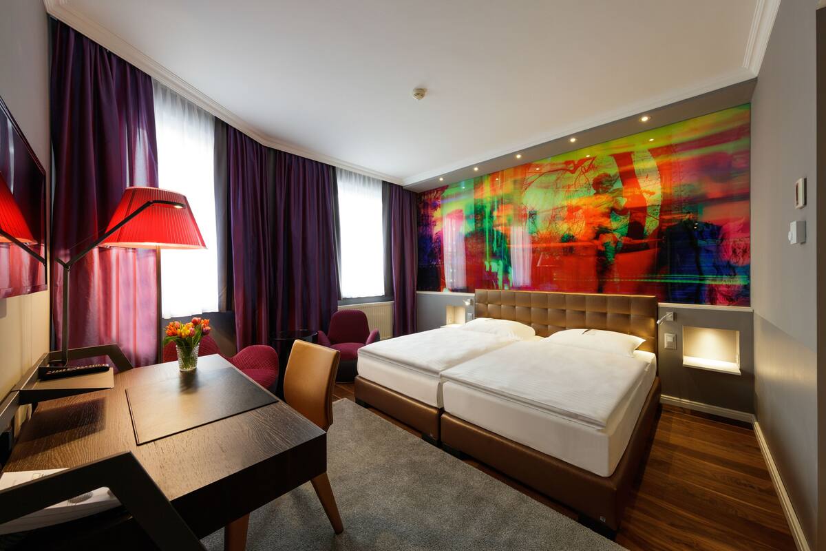 A hotel room with a bed, a desk, a chair, a lamp, and a colorful abstract wall art.