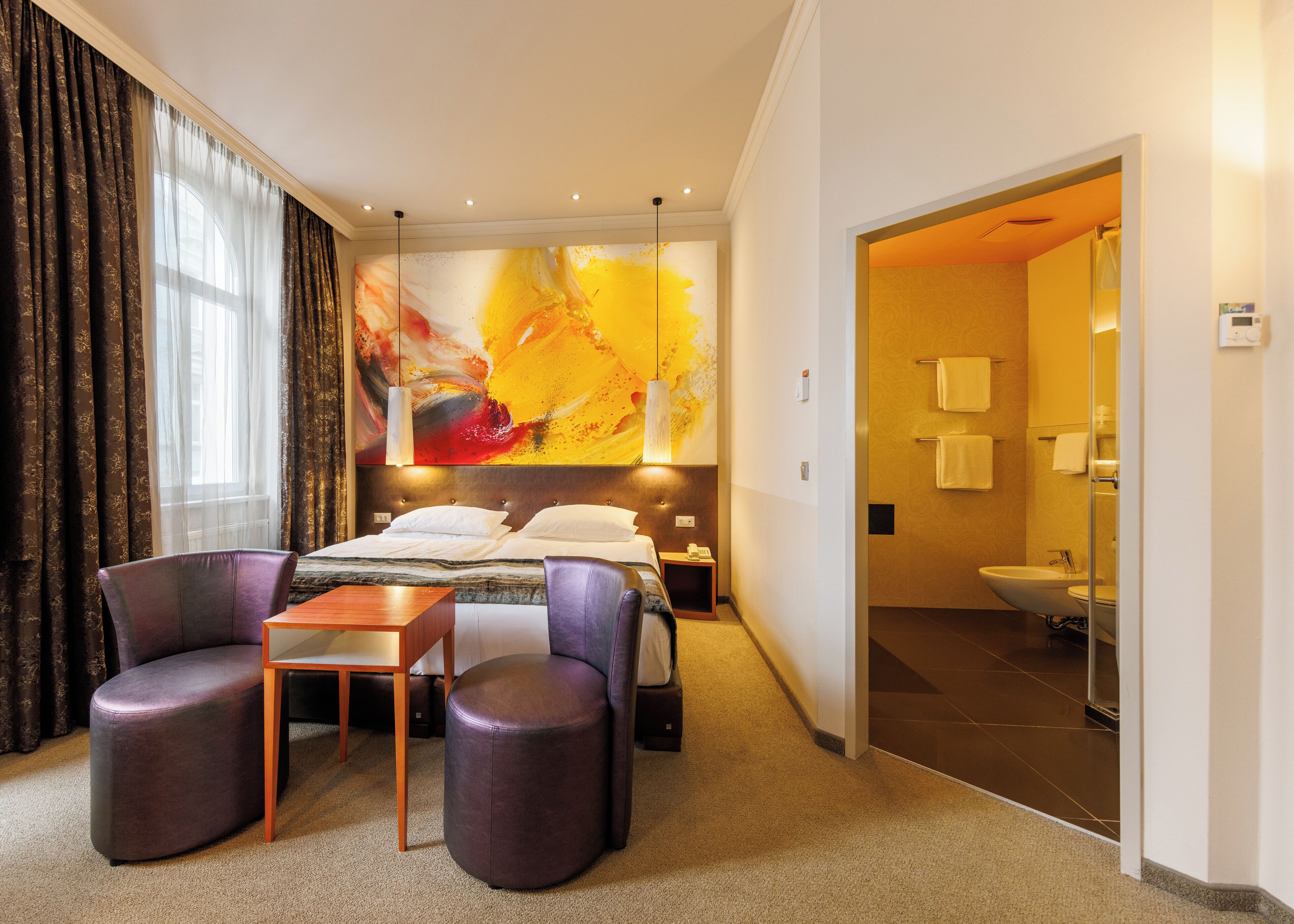 Une chambre d’hôtel avec un lit, deux fauteuils, une petite table et un grand tableau abstrait accroché au mur.
