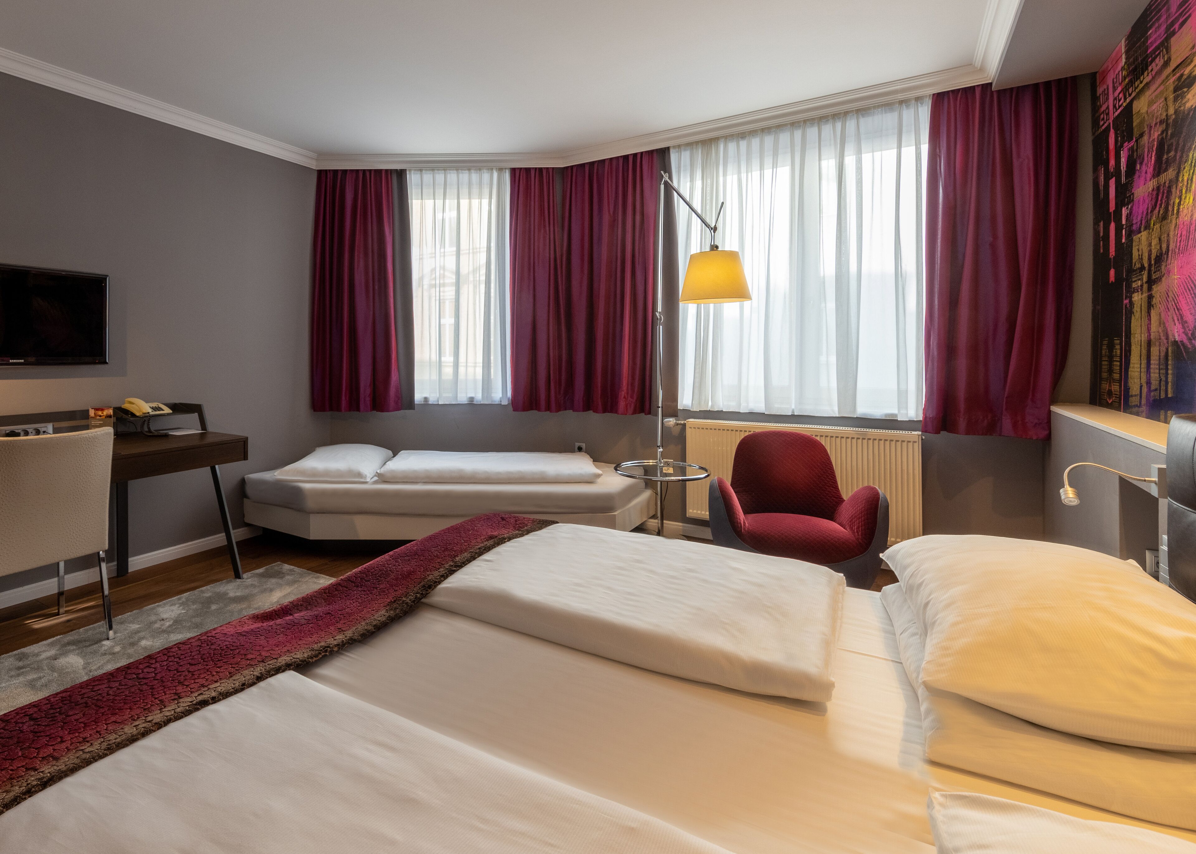 Quarto de hotel com duas camas, uma escrivaninha, uma cadeira, televisão e uma janela grande com cortinas.